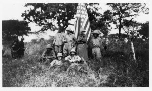 Former President Theodore Roosevelt (1858-1919) and other members of his expedition party from the Smithsonian Roosevelt African Expedition stand next to an American flag. Roosevelt is standing to the left of the flag with his head to the side. Other men in the image include Kermit Roosevelt, Edgar Alexander Mearns, and John Alden Loring. On this trip, Roosevelt collected natural history specimens for the United States National Museum (now National Museum of Natural History) and live animals for the National Zoological Park